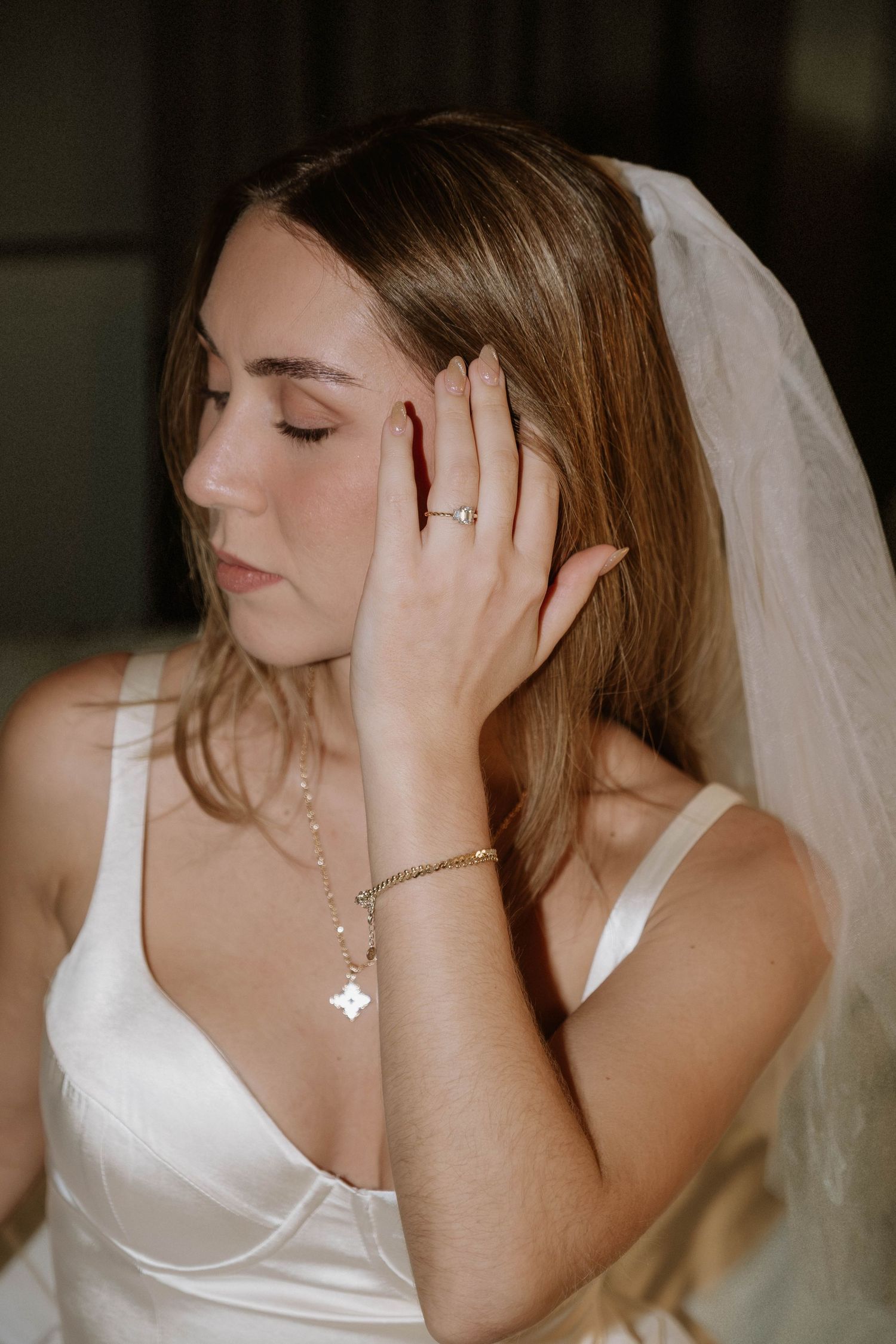 A bride in a white wedding dress touches her face gently while wearing a delicate necklace and veil.