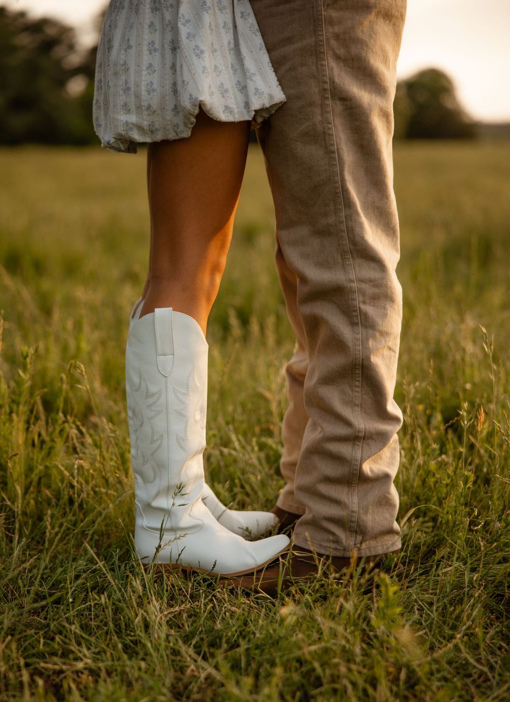 Two pairs of boots stand close together in a grassy field at sunset, one white cowboy boots and one brown pants and boots.
