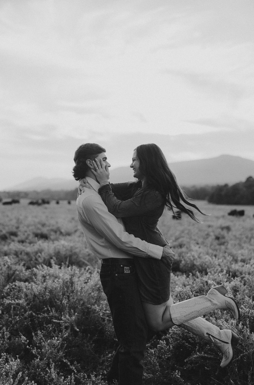 A couple embraces in a scenic mountain field during sunset, captured in black and white.