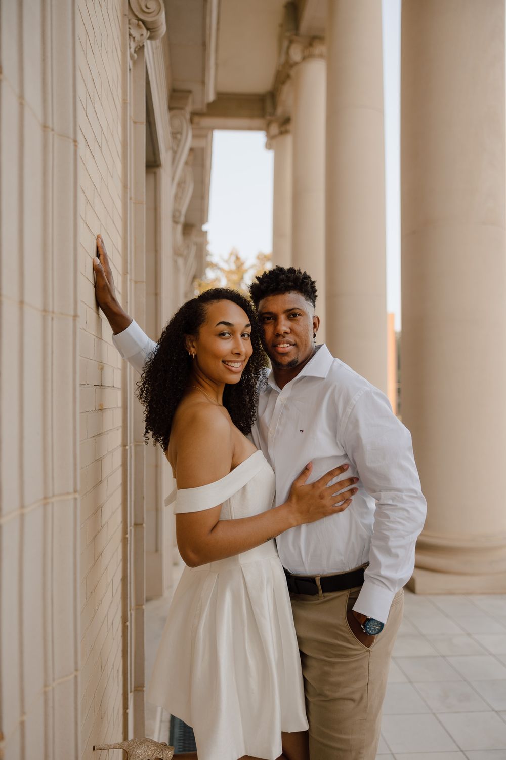 A couple shares joyful moments in an elegant colonnade setting wearing coordinated white and beige outfits.