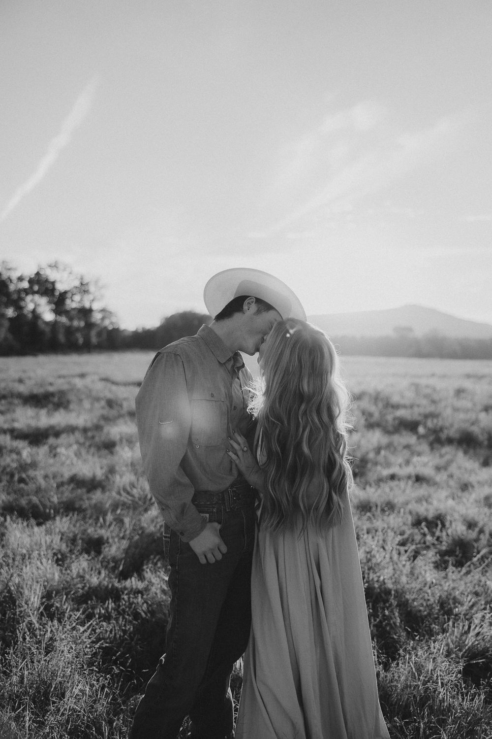 A couple in western attire shares a tender moment in a scenic field at sunset.