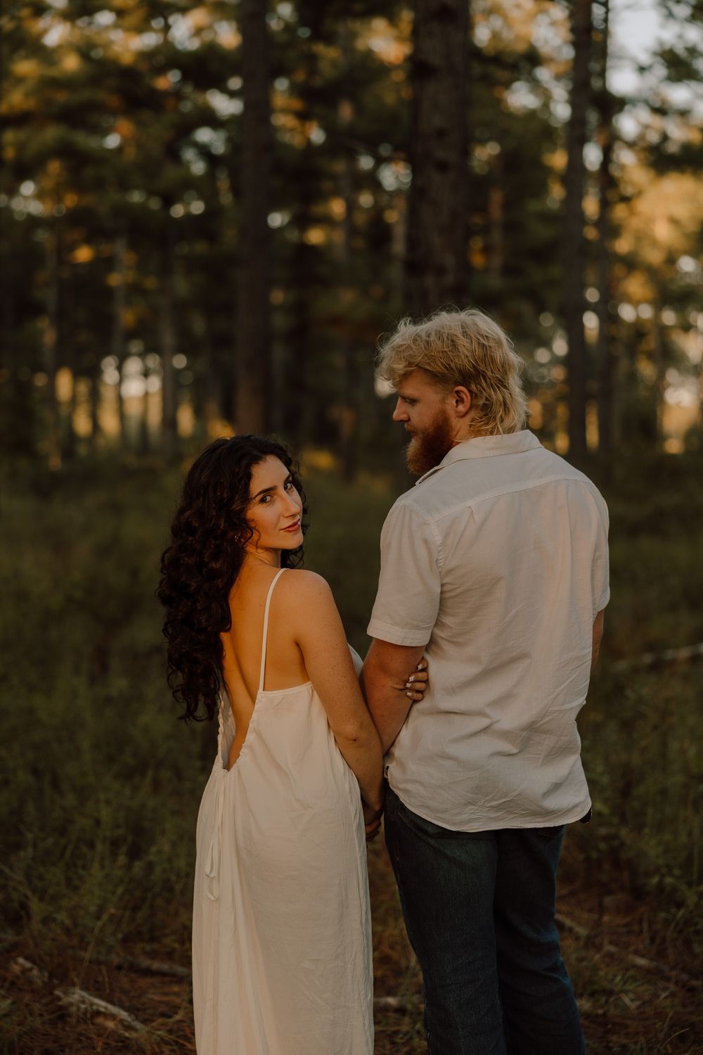 A couple in white clothing share an intimate moment while walking through a sunlit forest at golden hour.