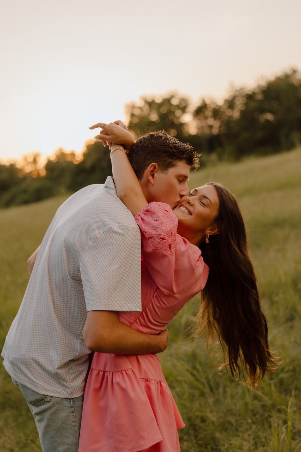 A couple embraces in a grassy field at sunset wearing white and coral pink outfits.