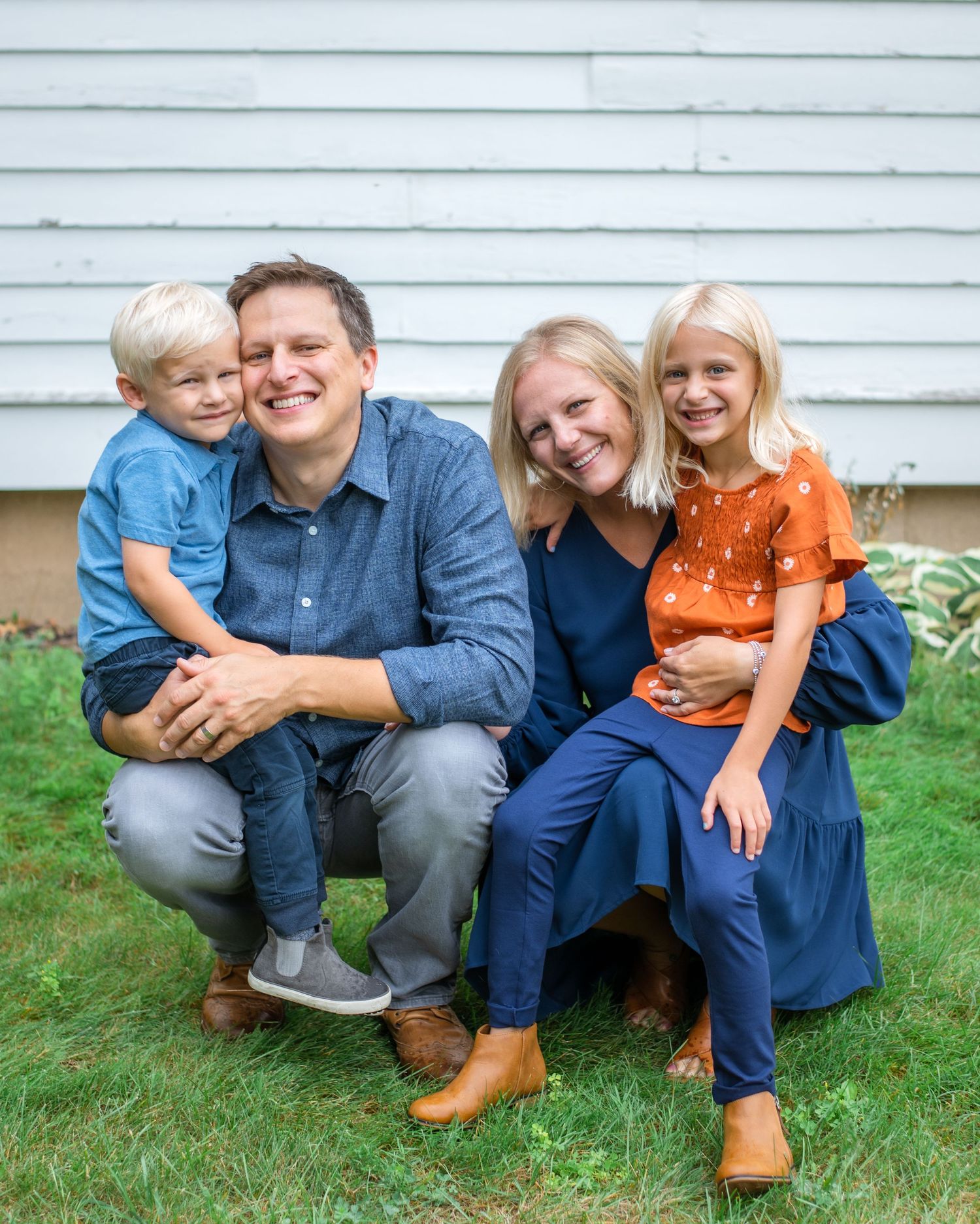 A family poses together outdoors in front of a blue siding wall while sitting on grass in coordinated casual outfits.