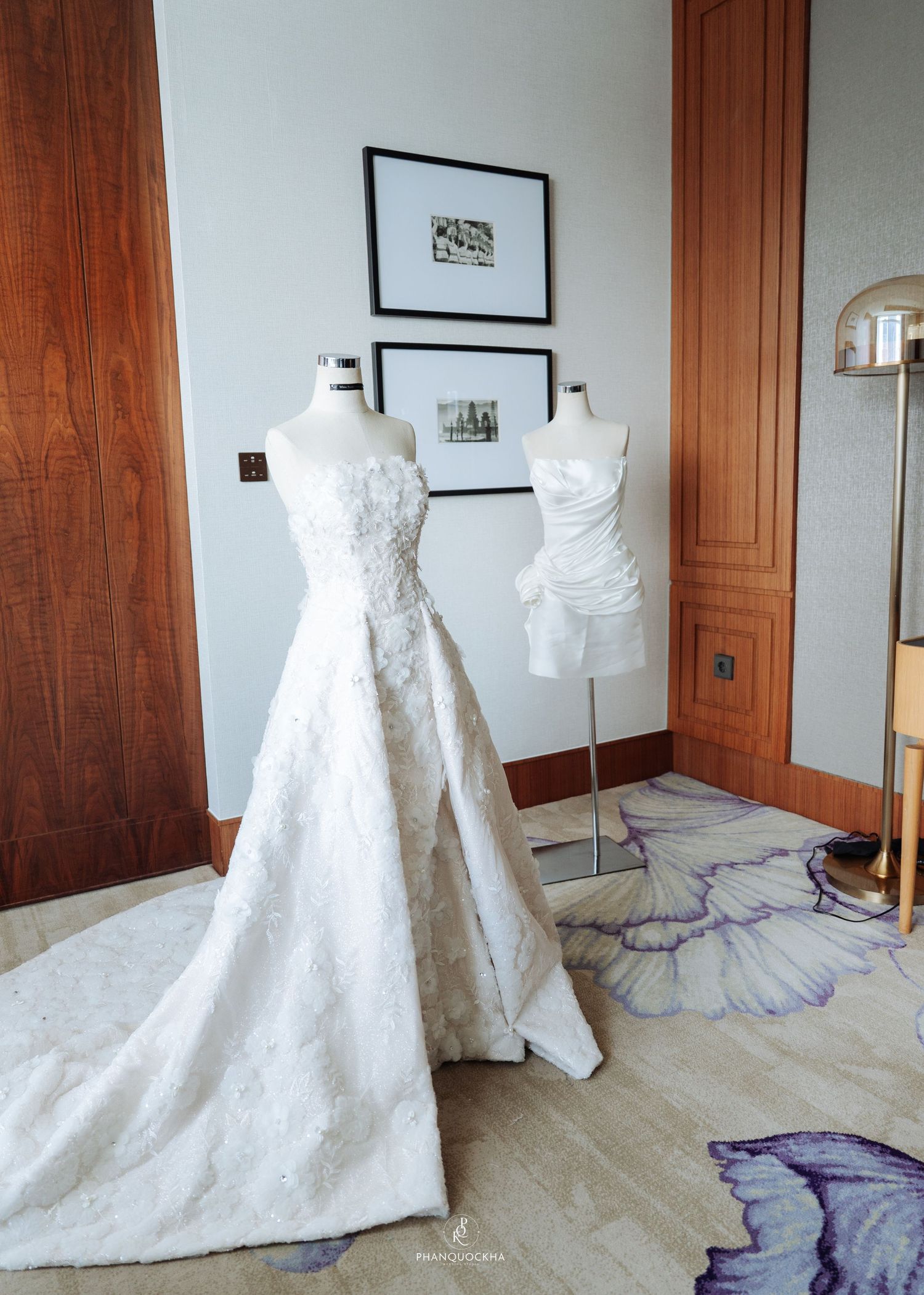 Two elegant strapless wedding gowns displayed on mannequins in a luxury hotel suite with floral carpet.