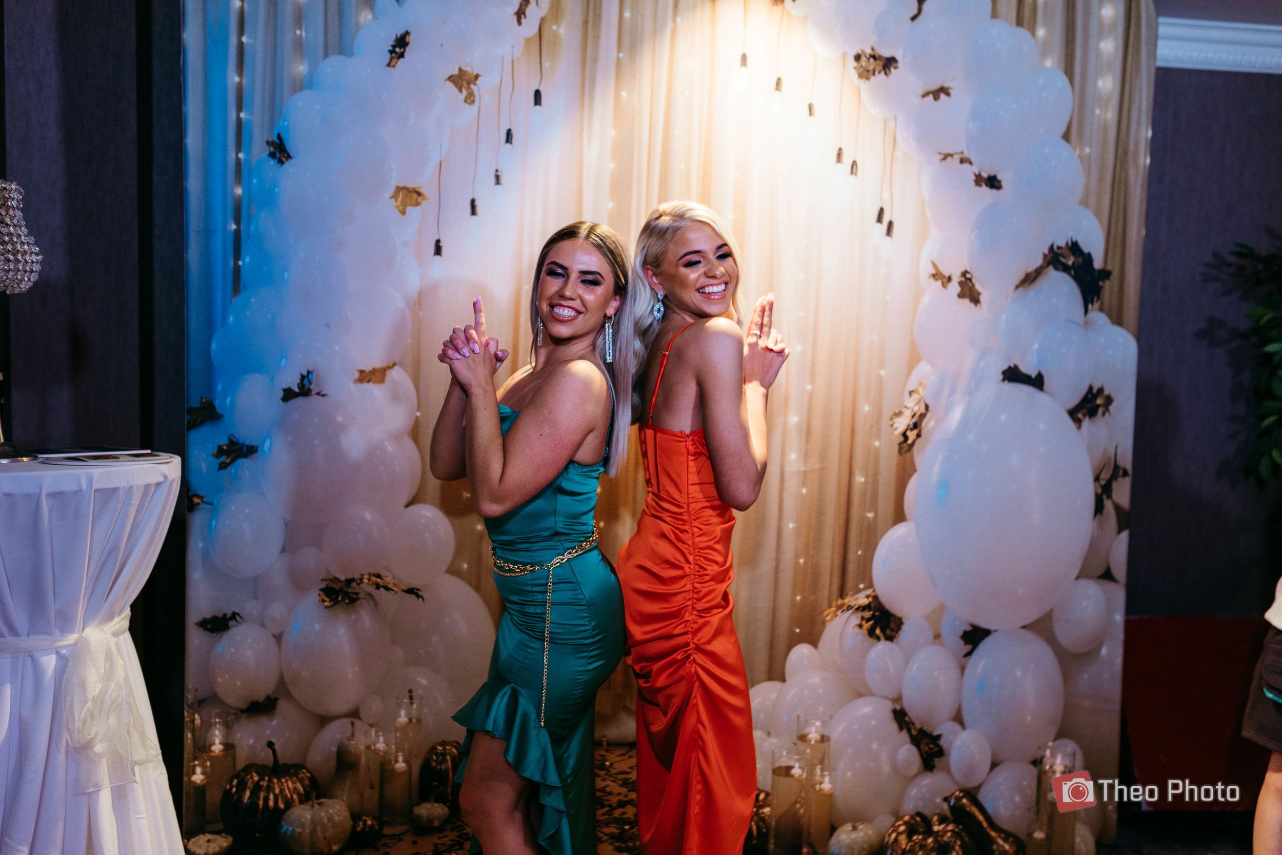 Two people in colorful dresses dance and pose together in front of a butterfly-decorated backdrop at an event.