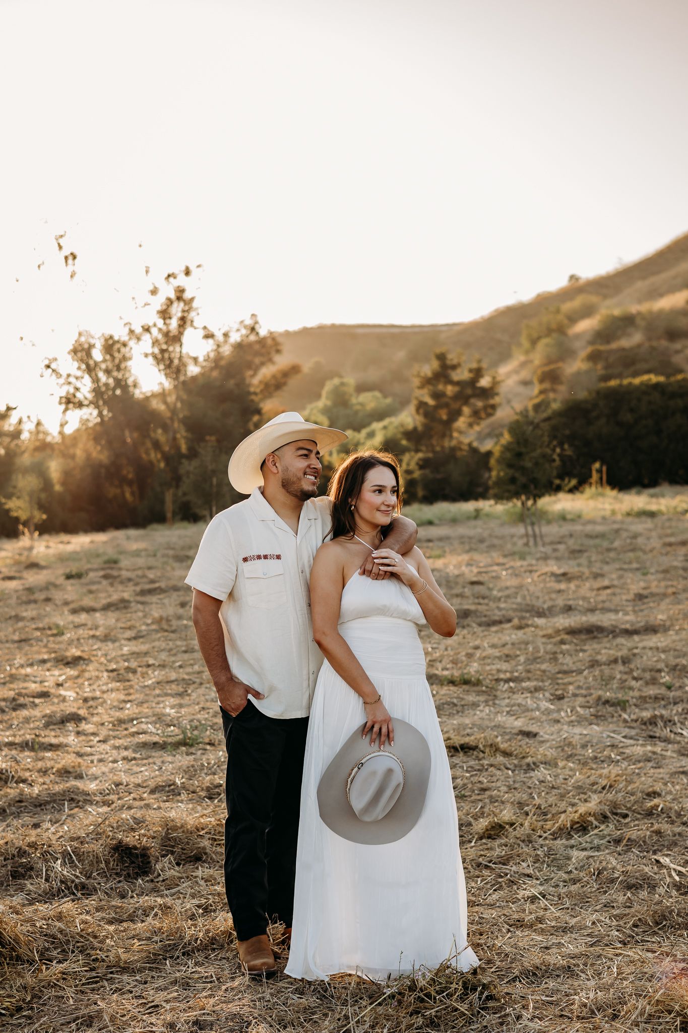 Couple in white outfits and cowboy hats posing in a golden field at sunset with rolling hills behind them.