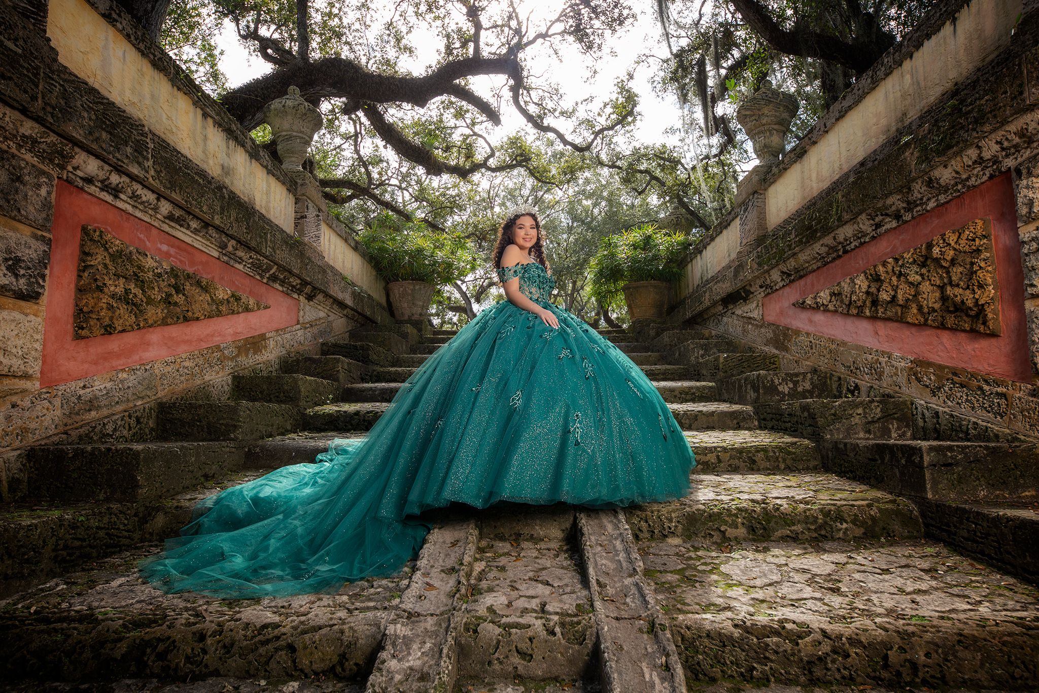 A stunning emerald green quinceañera gown with a flowing train photographed on ancient stone steps in a garden setting.