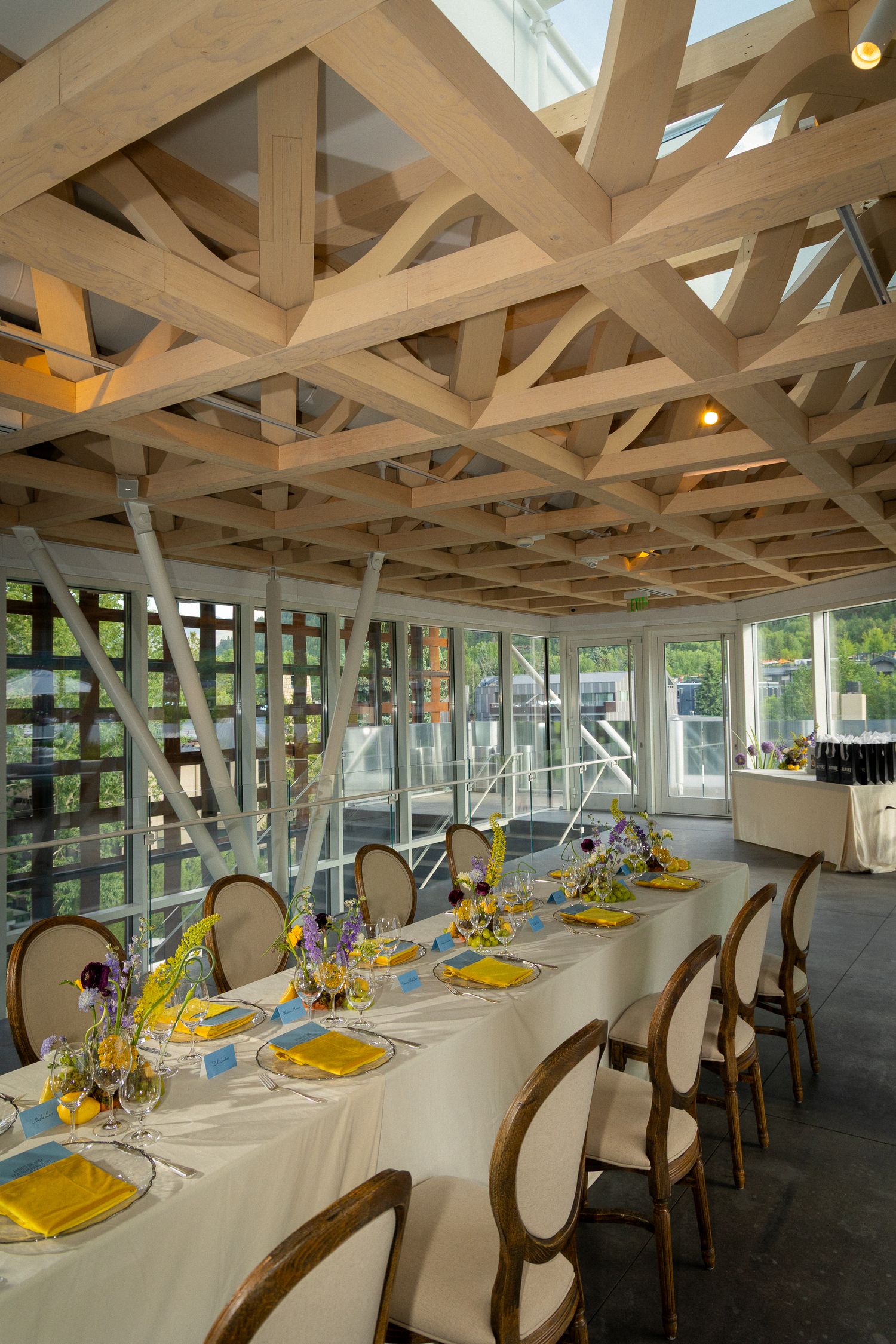 Long banquet table set with yellow placemats and floral centerpieces in a bright sunlit room with exposed wooden beams.