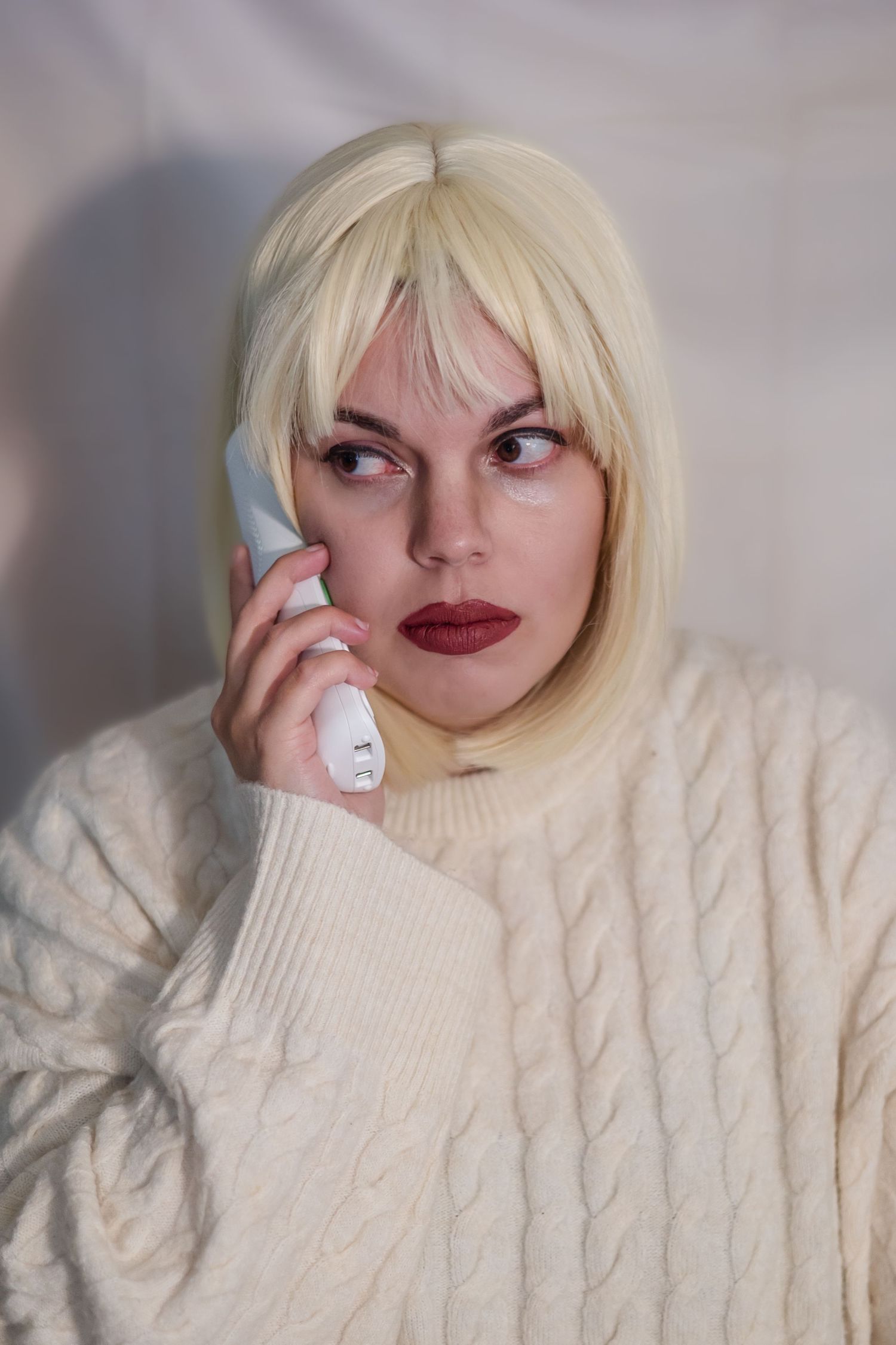 A person wearing a cream cable knit sweater poses playfully with blonde bangs and red lipstick in a photo series.