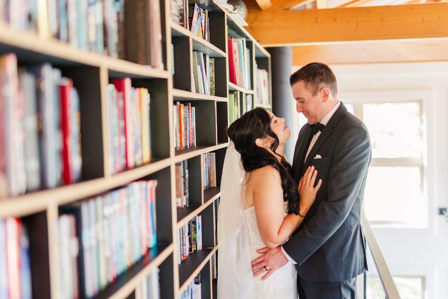 A couple shares a romantic moment between bookshelves in a library setting.
