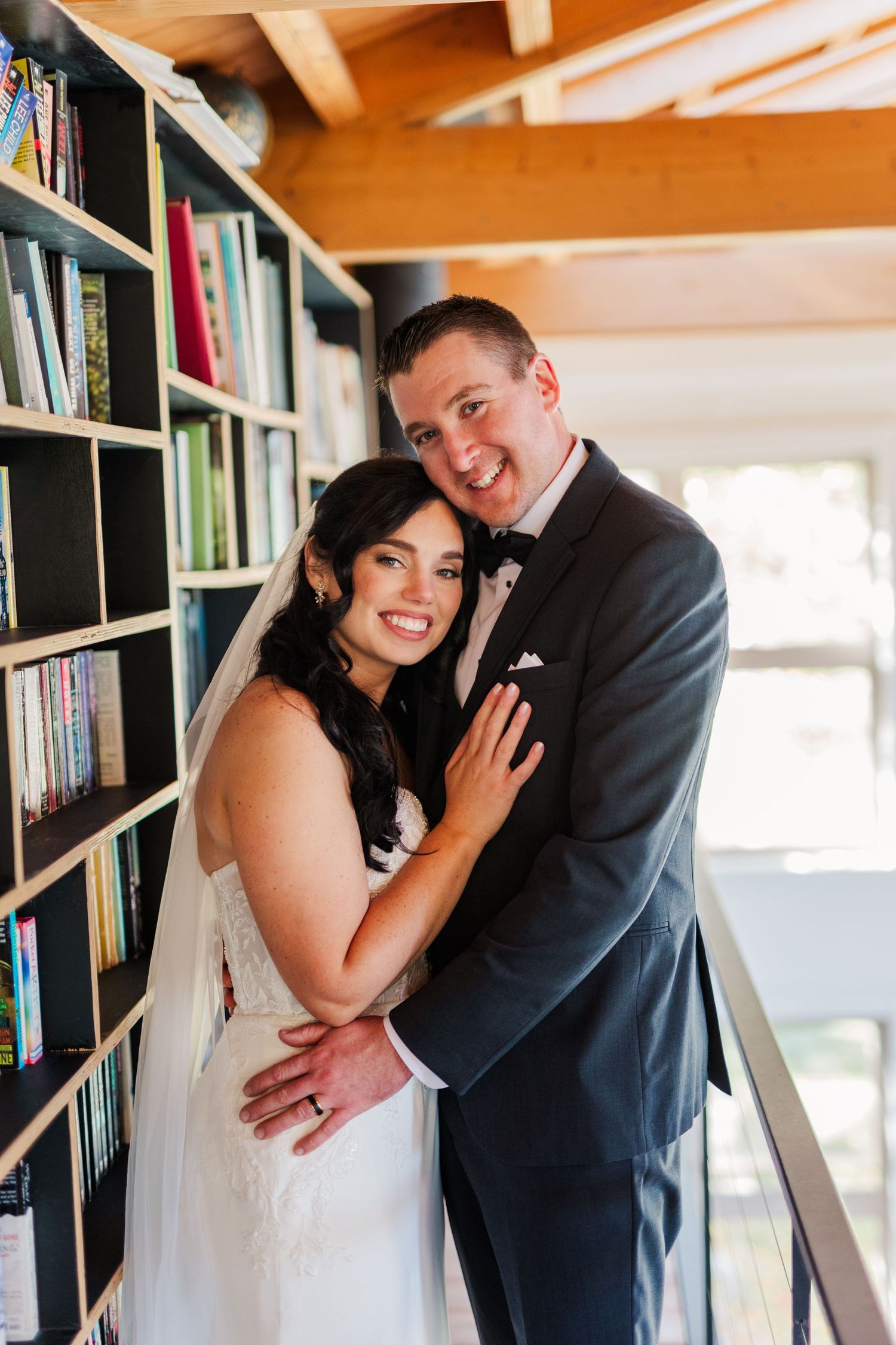 A romantic wedding couple shares intimate moments while posing against a bookshelf in a bright indoor setting.
