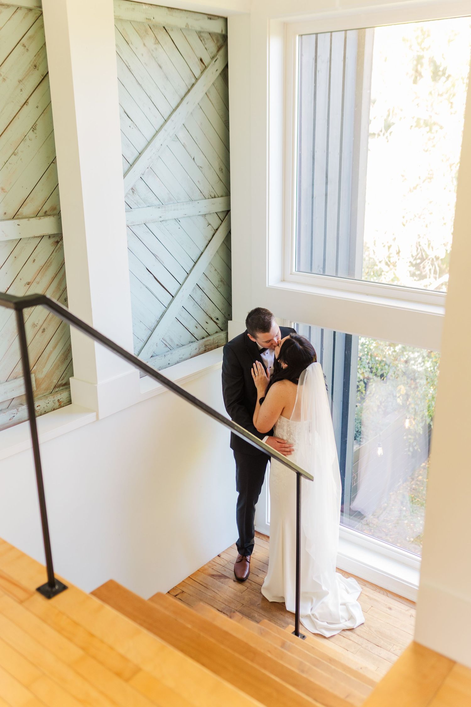 A couple shares a romantic moment in a bright white stairwell with wooden floors and diagonal wall paneling.