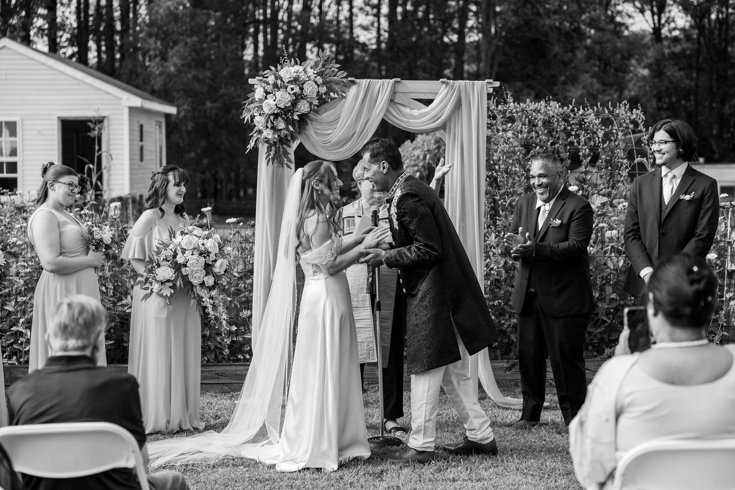 A black and white wedding ceremony photo showing an outdoor garden wedding with guests and decorations.