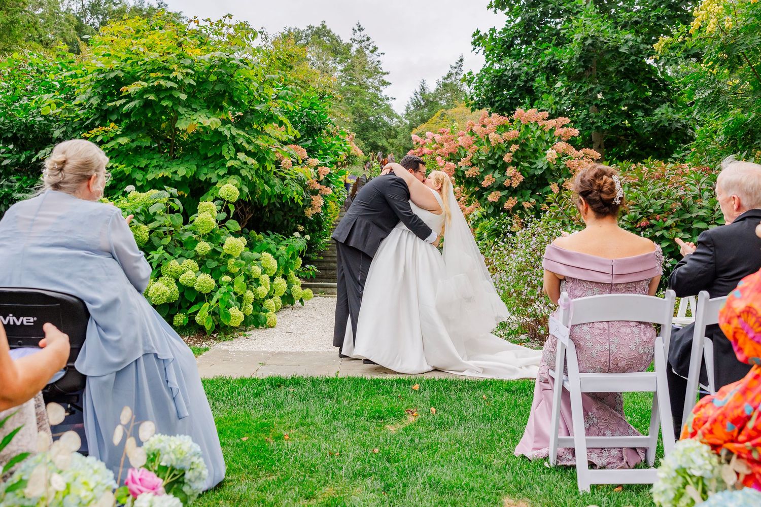 Wedding guests gather in a lush garden setting with vibrant green foliage and blooming flowers during an outdoor celebration.