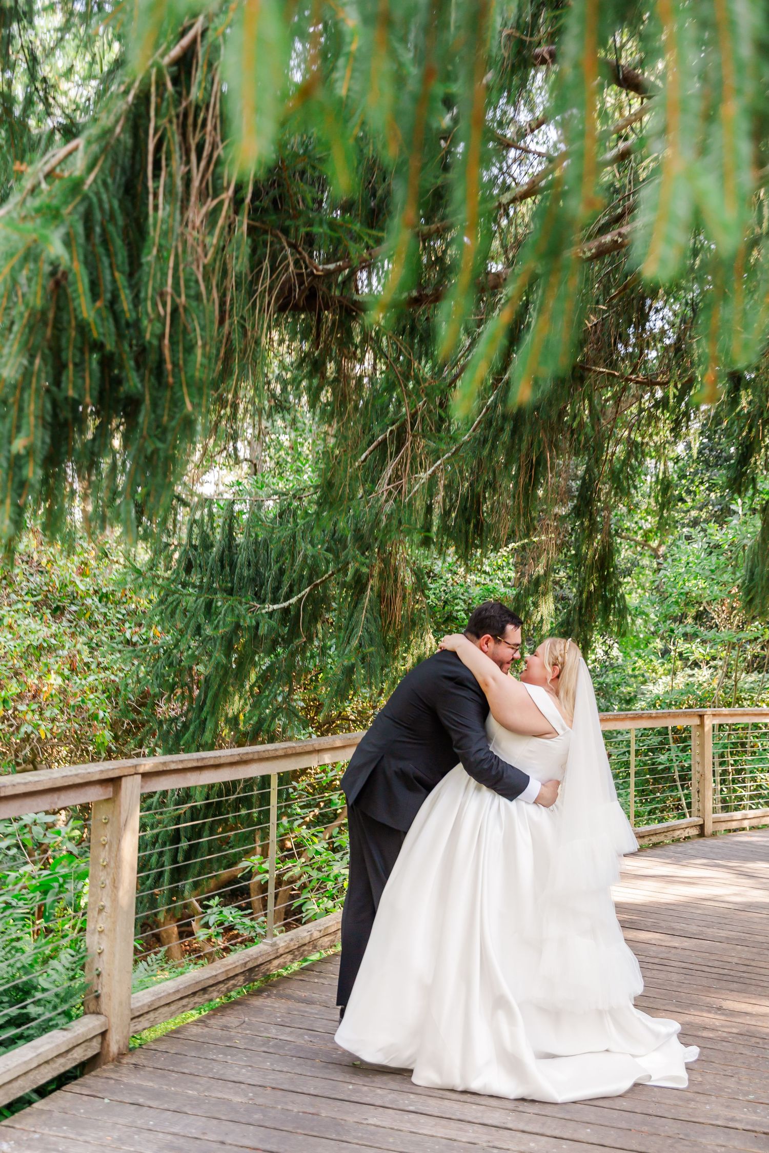 A couple in formal attire shares a romantic embrace on a wooden boardwalk surrounded by lush greenery.