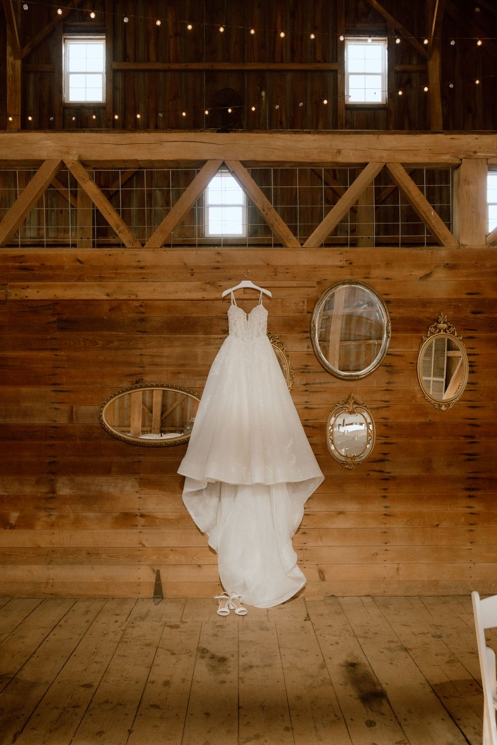 A white lace wedding gown hangs on a wooden barn wall surrounded by vintage oval mirrors and string lights above.