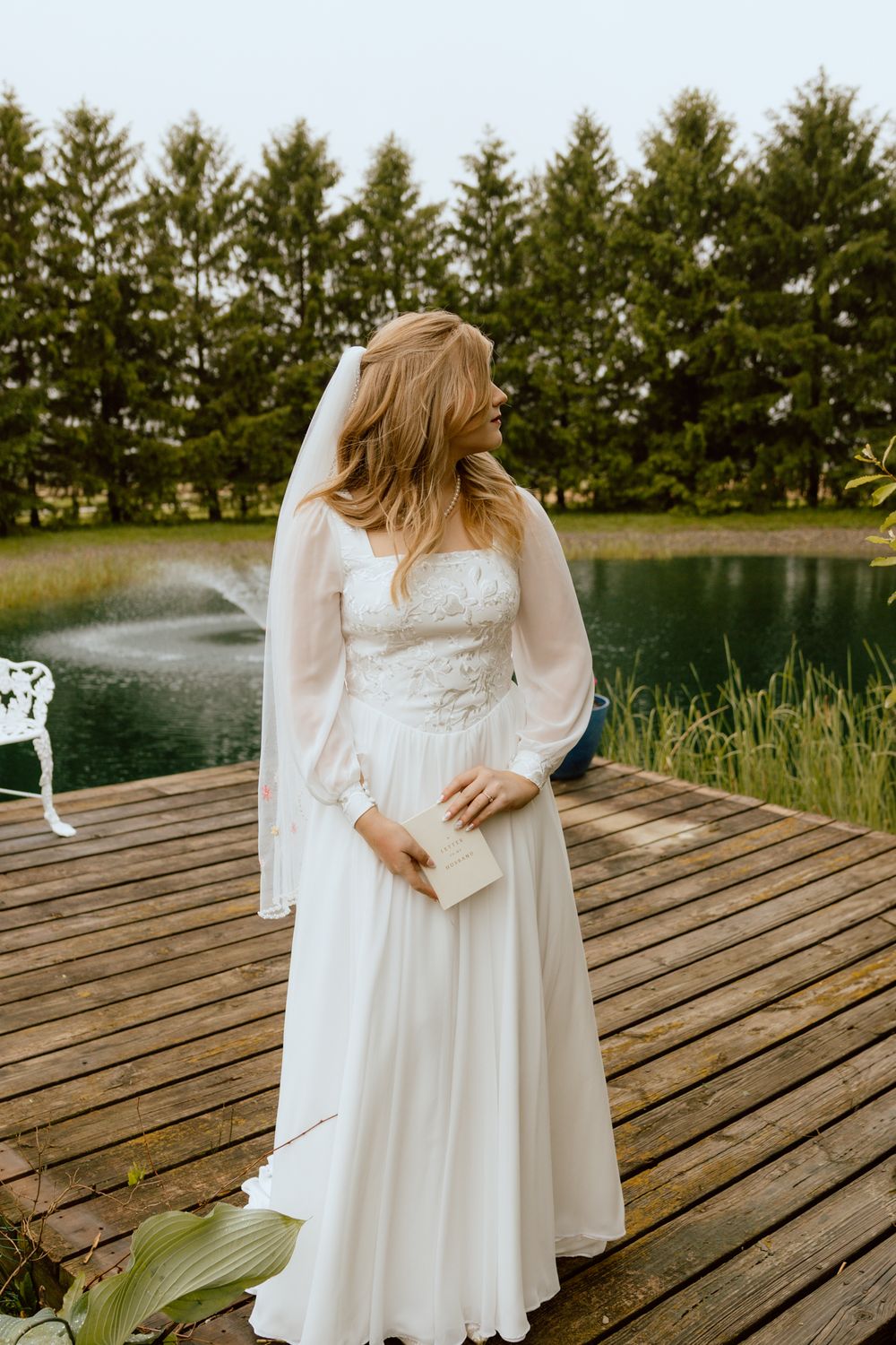 Elegant bride in a long-sleeve lace wedding gown holding a book on a wooden dock near a pond.