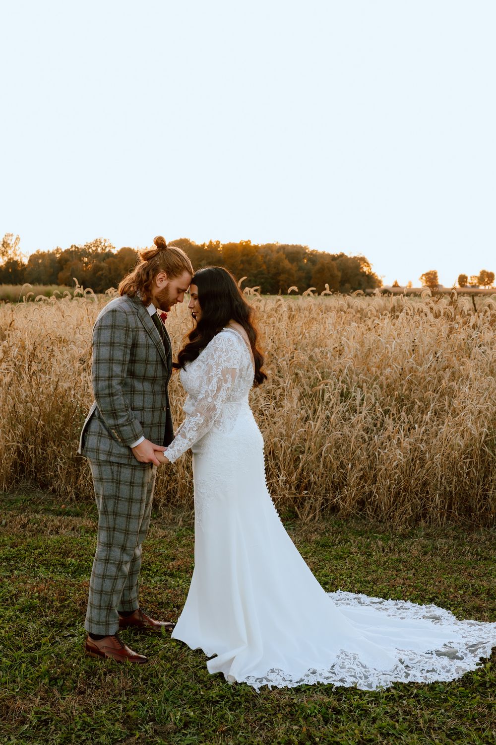 Bride and groom touching foreheads tenderly in front of a golden wheat field at sunset.