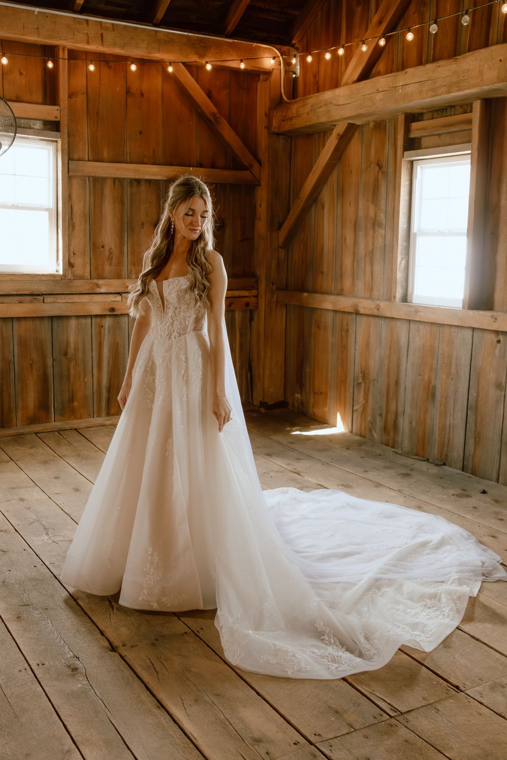 A bride in a strapless lace ball gown stands gracefully inside a rustic wooden barn with warm natural light.