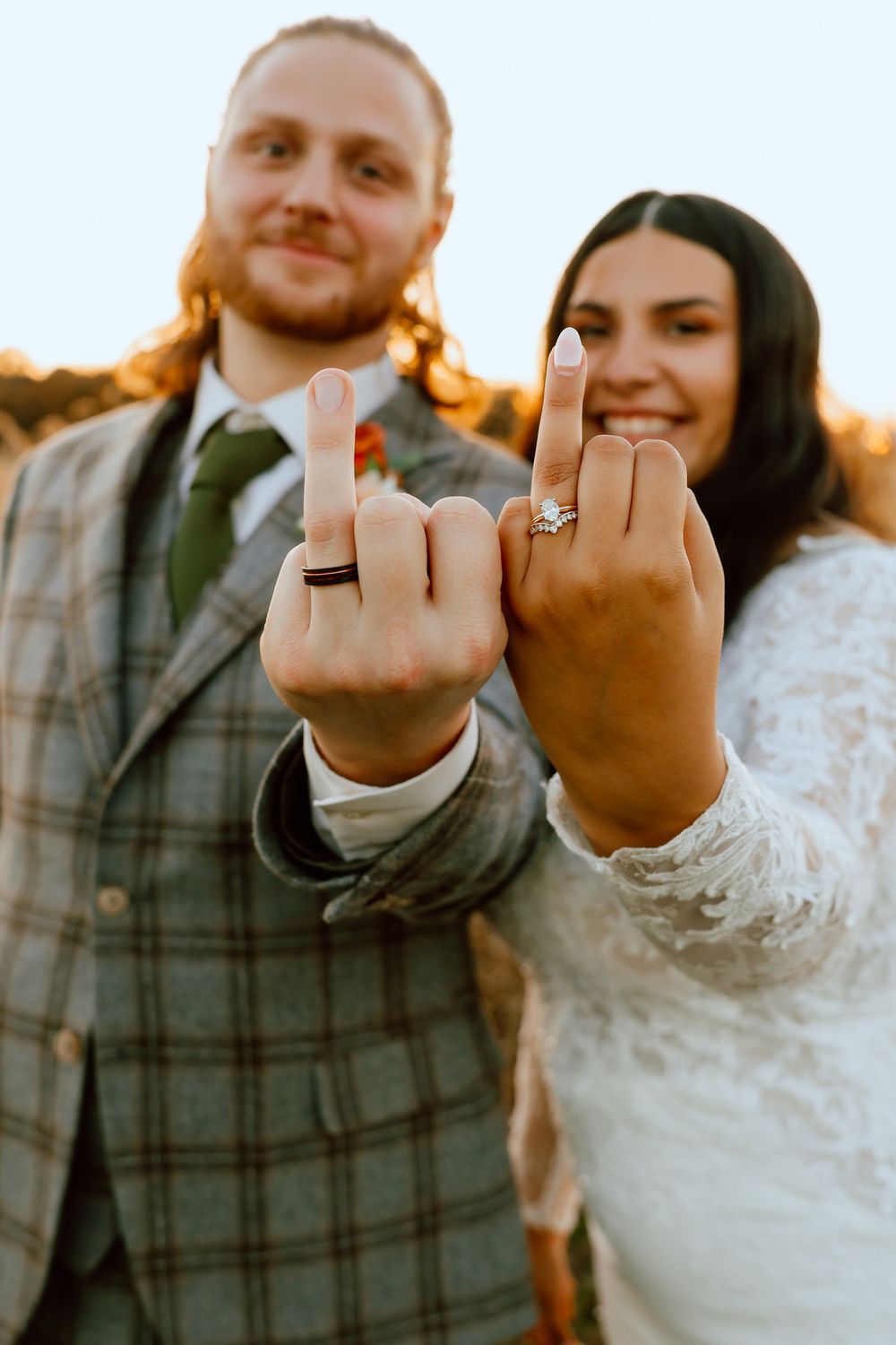 Newlyweds proudly showing off their wedding rings with a playful gesture at golden hour outdoors.
