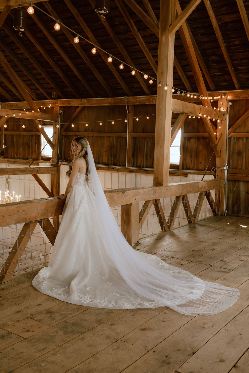 A bride in a cathedral-length veil and lace gown stands inside a rustic barn adorned with warm string lights.
