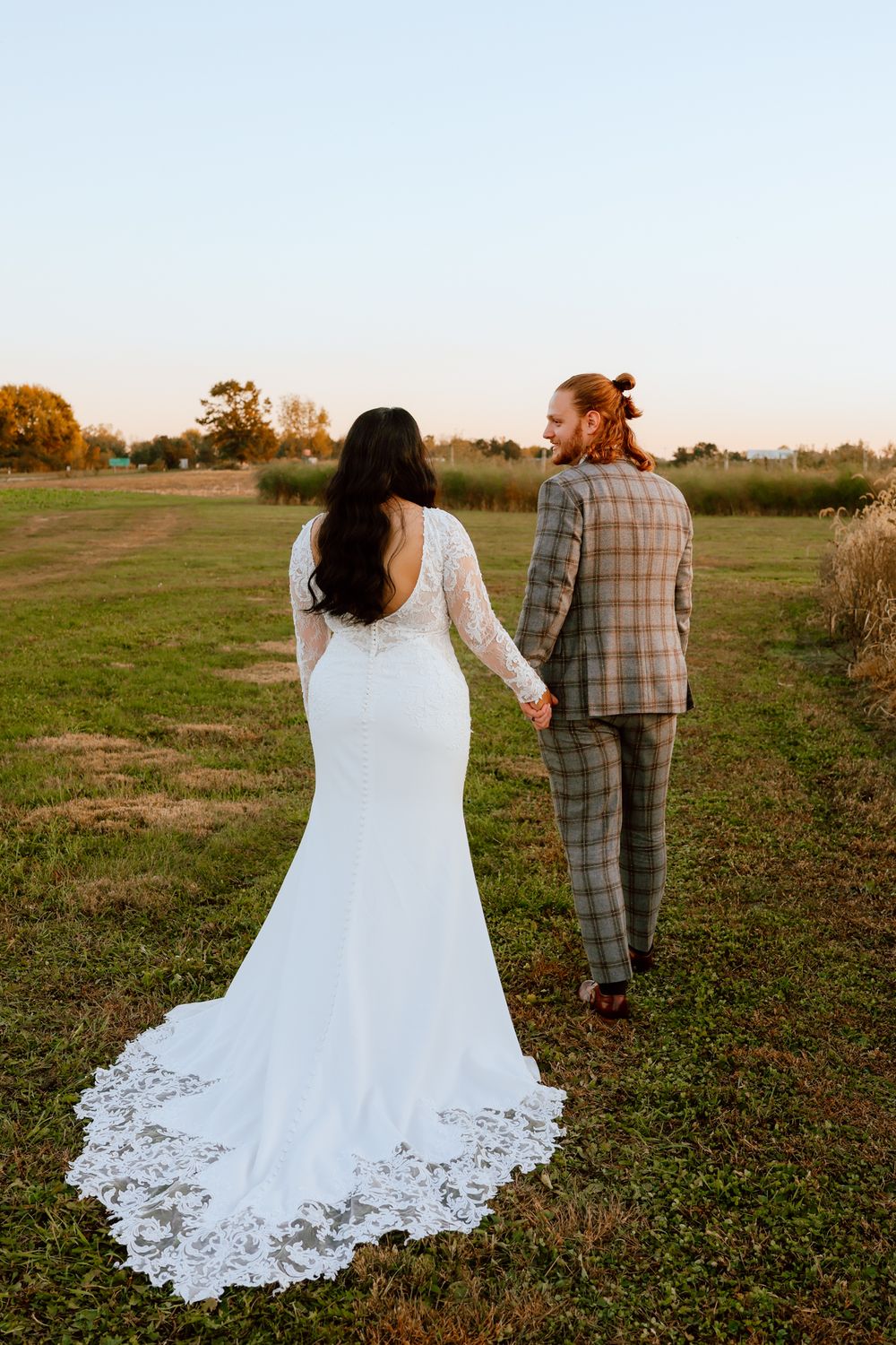 Bride in a lace mermaid gown and groom in plaid suit holding hands walking through a golden field.