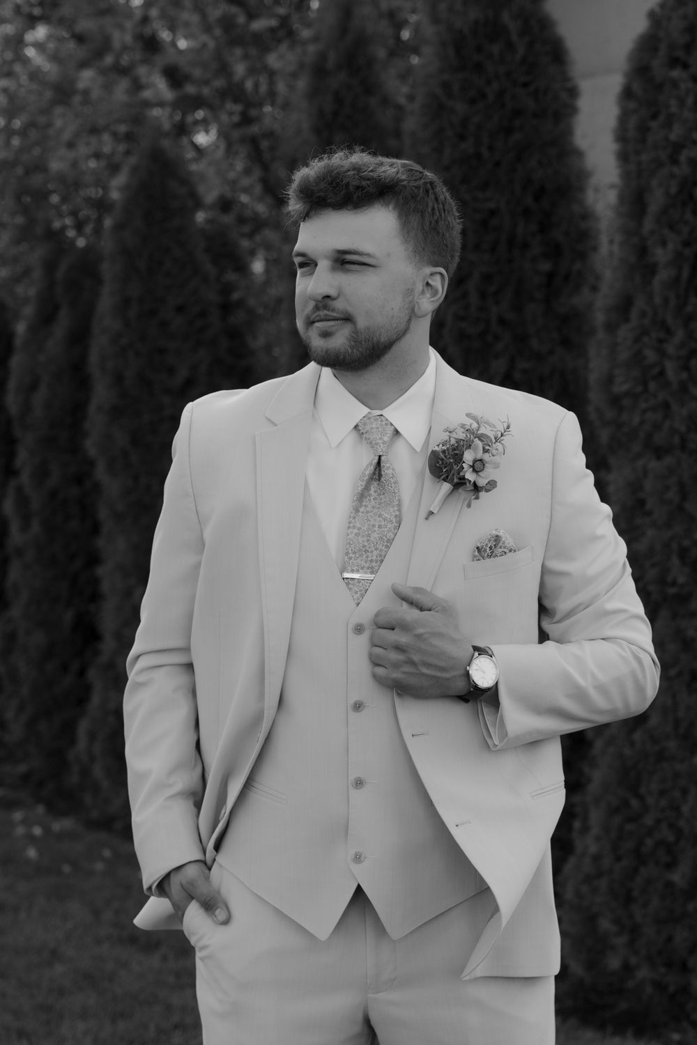 A dapper groom in a light beige three-piece suit with floral boutonniere poses confidently in a black and white portrait.
