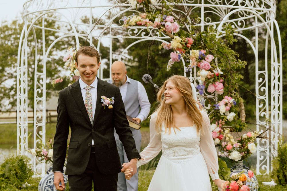 Happy bride and groom walk hand in hand beneath floral arch after ceremony.