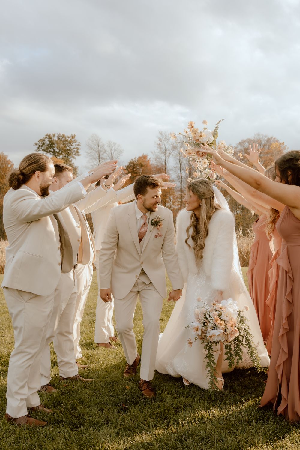 A bride and groom walk through an arch of raised hands formed by their wedding party on an autumn lawn.