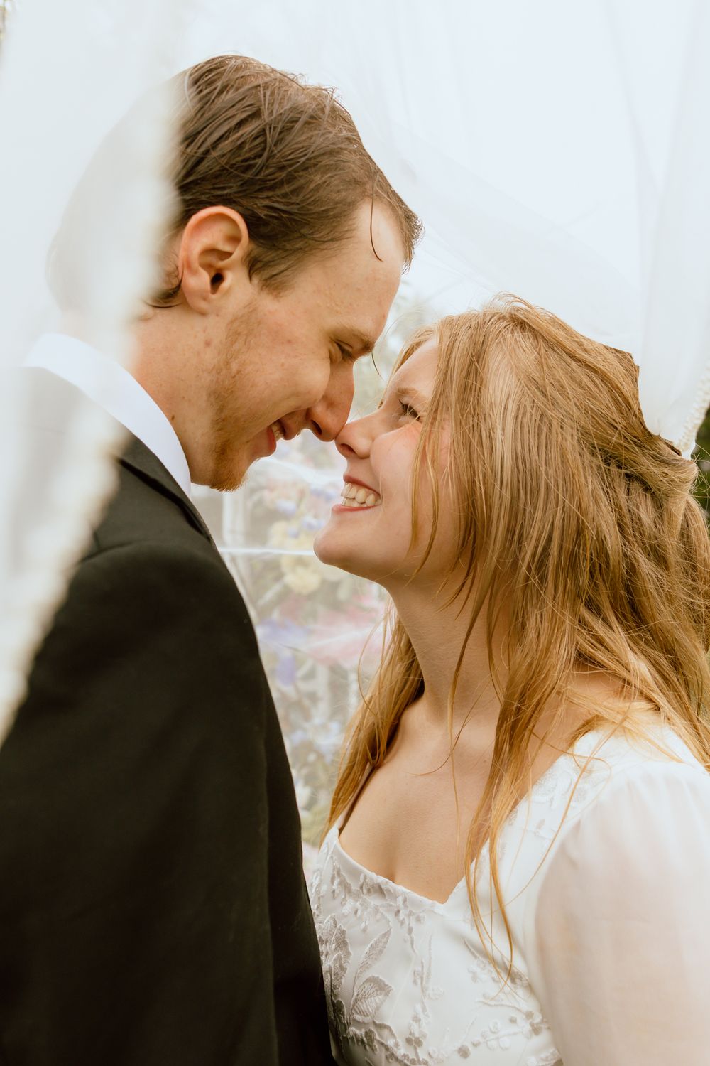 Smiling bride and groom touch foreheads tenderly beneath flowing wedding veil.