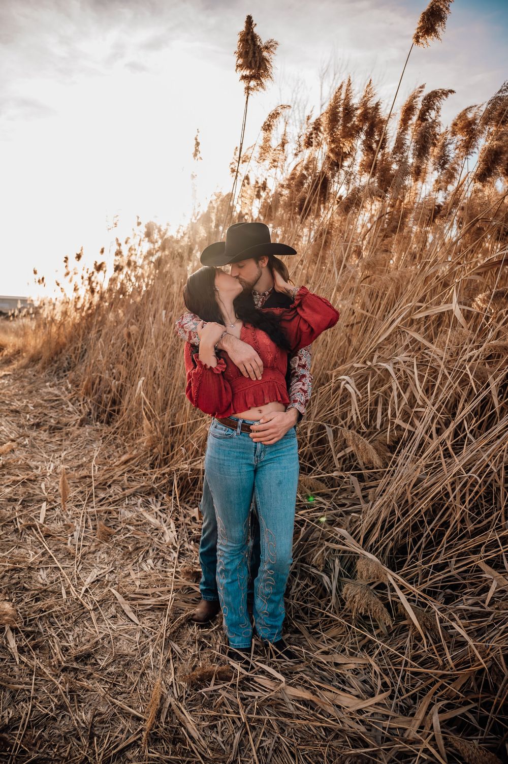 Couple embracing in rustic field with dried grass and moody sky backdrop.