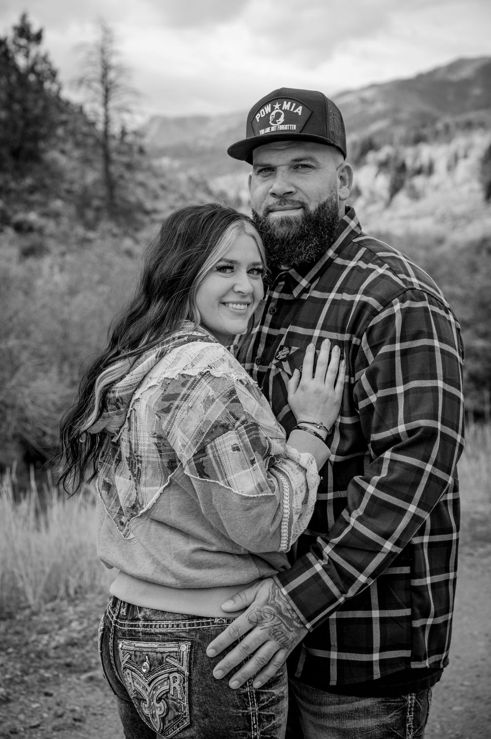 A couple in matching plaid shirts pose for a black and white portrait against a mountain landscape.