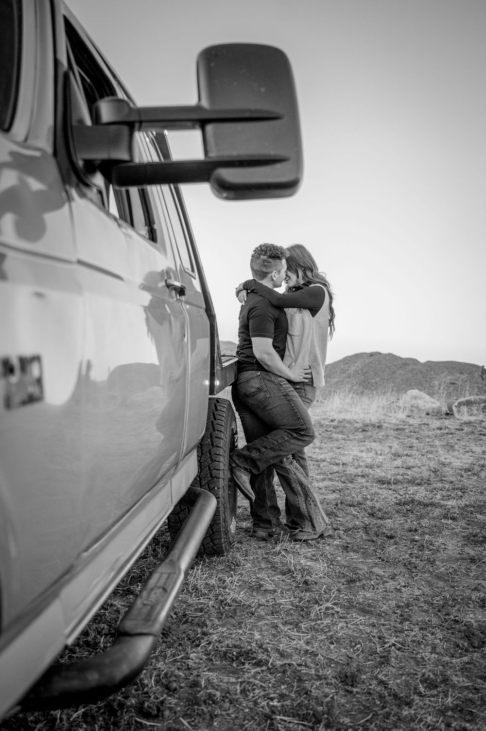 Black and white photo of a couple embracing next to a truck in a rural setting.