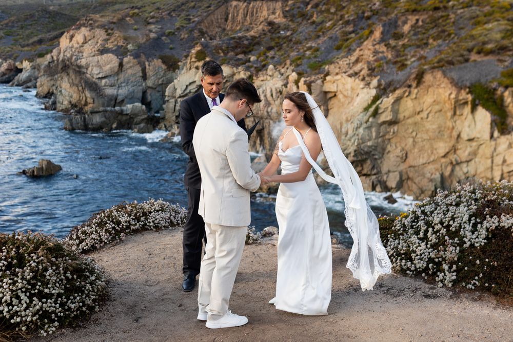 Couple exchanging vows at a cliffside wedding ceremony overlooking the ocean with rocky coastline backdrop.