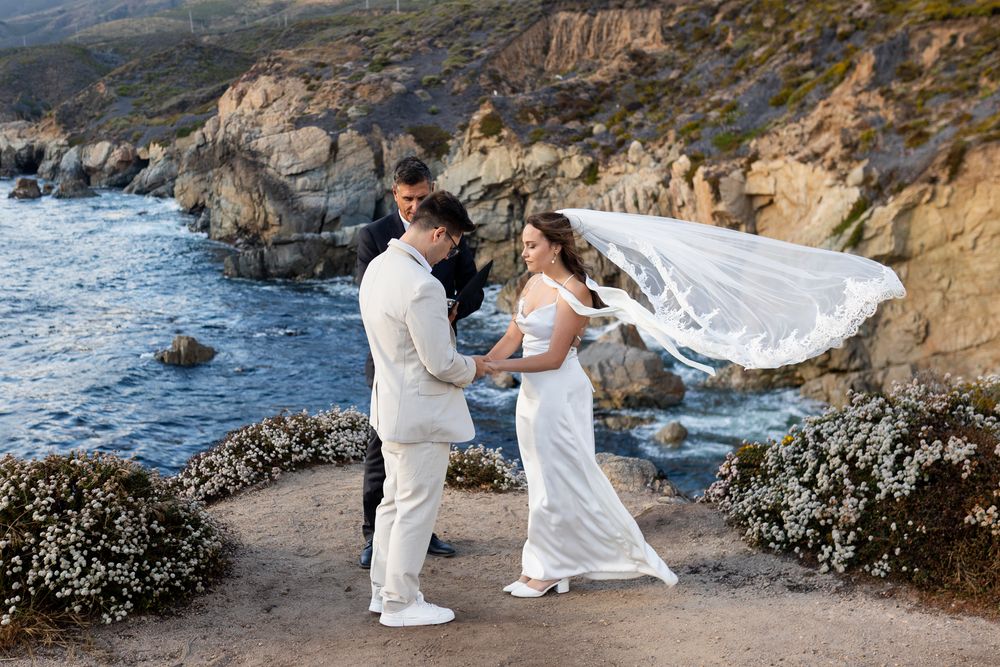 Couple in white attire poses along rocky coastal cliffs with ocean waves crashing in the background.