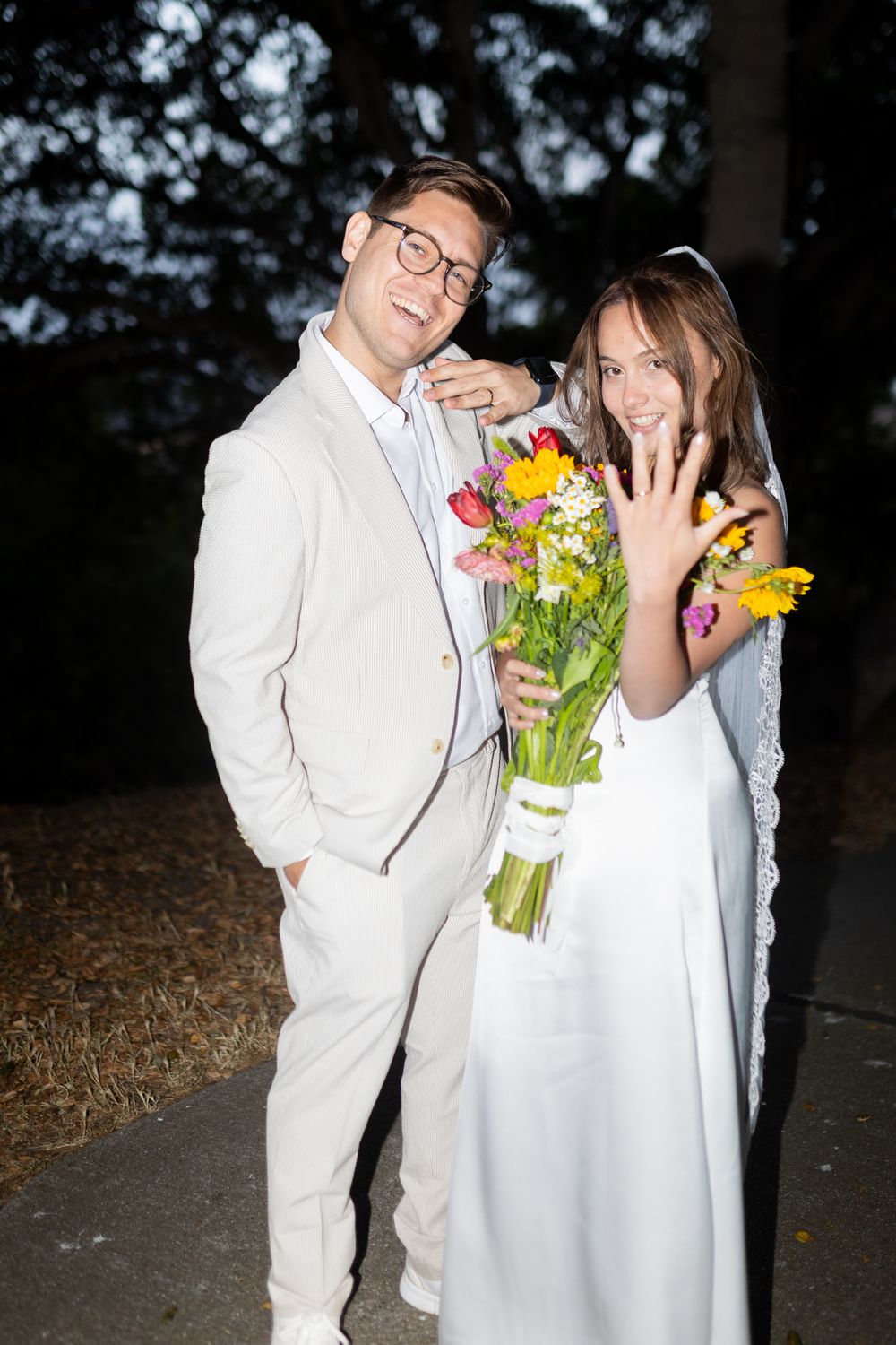 A couple in white attire shares playful moments while holding a vibrant bouquet of flowers at dusk.