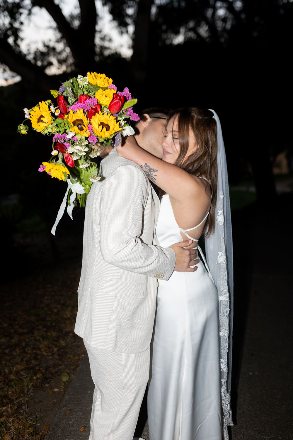A couple in white wedding attire share a kiss while holding a vibrant yellow and pink floral bouquet in a dark outdoor setting.