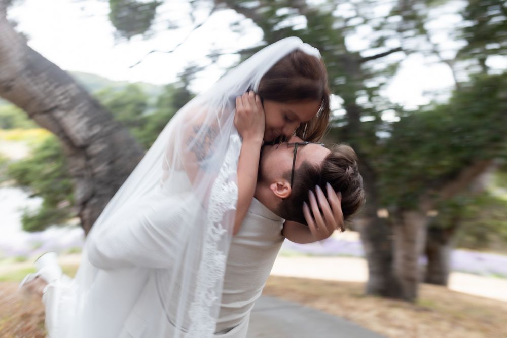 A romantic wedding couple embraces in an outdoor garden setting with soft natural lighting and blurred greenery background.