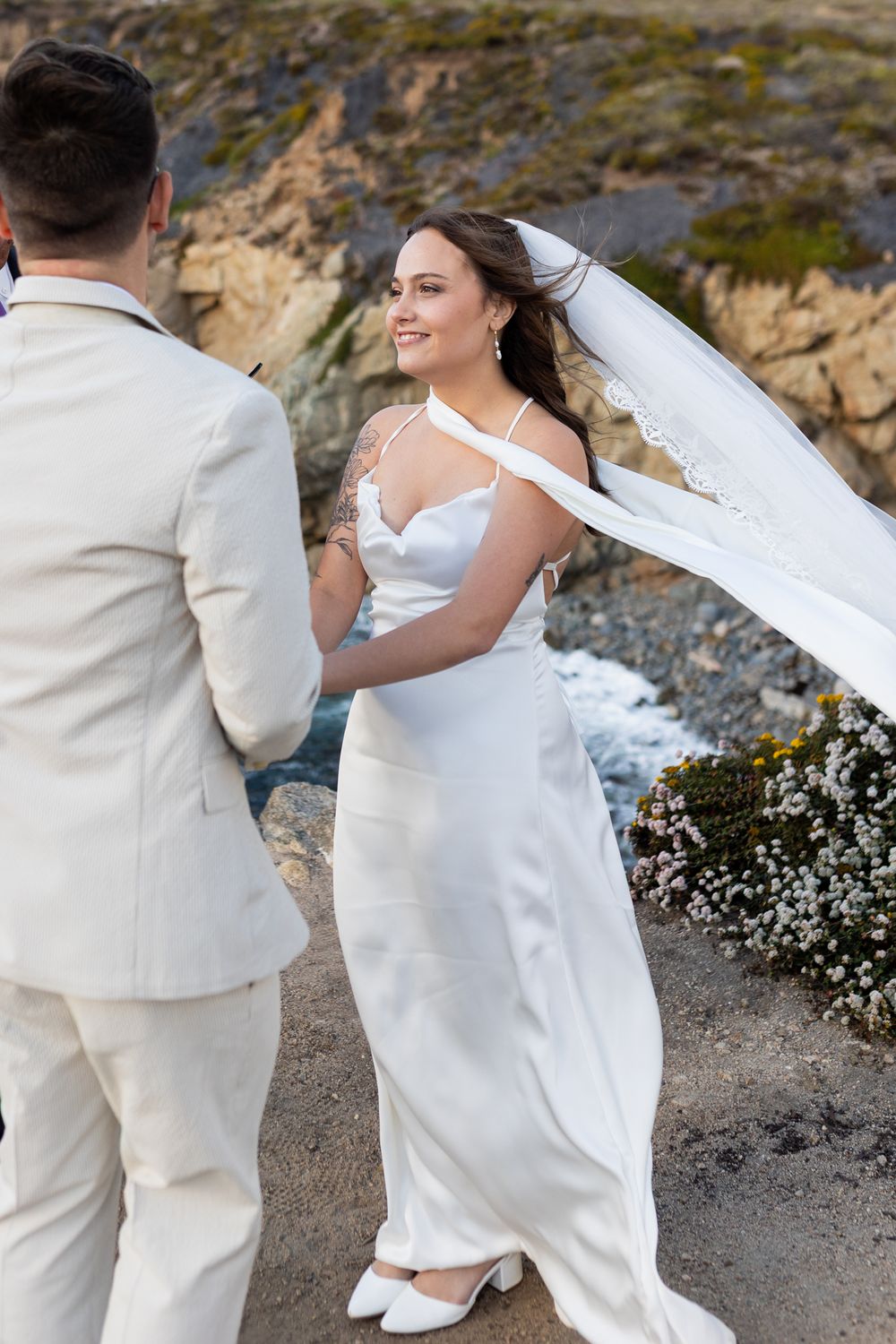A couple in white attire dances together on a cliff edge with dramatic coastal scenery in the background.