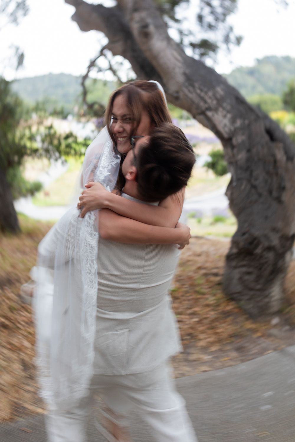 Two people share a tender embrace outdoors under a tree canopy with a flowing white dress creating movement.