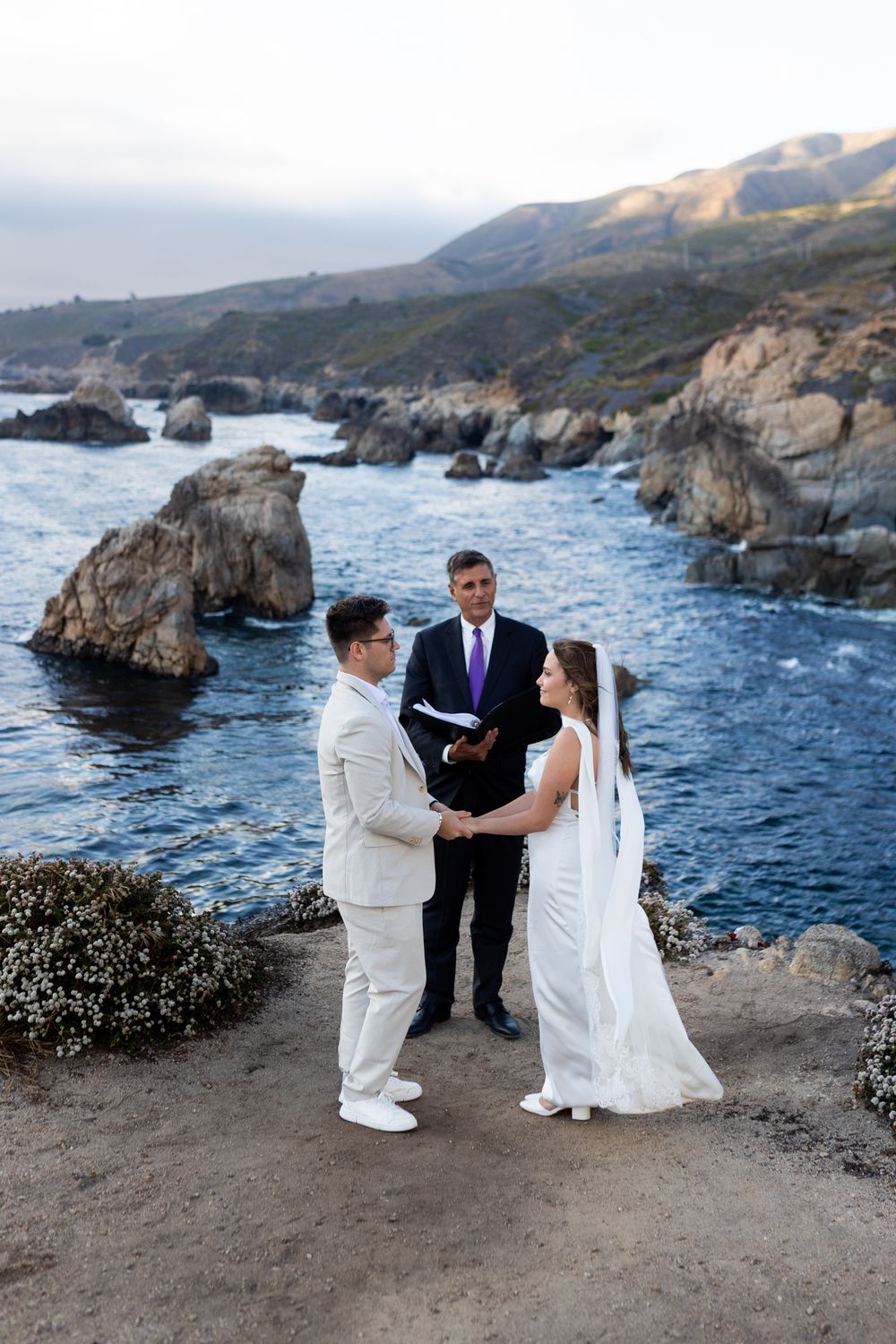 Couple exchanging vows at a coastal wedding ceremony with rocky cliffs and blue ocean in Big Sur, California.