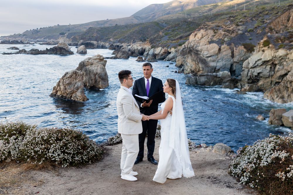 Couple exchanges wedding vows with officiant on rocky Big Sur coastline overlooking Pacific Ocean at sunset.