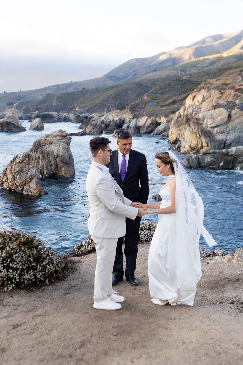 Couple exchanges vows at outdoor coastal wedding ceremony with rocky cliffs and ocean backdrop at Big Sur, California.