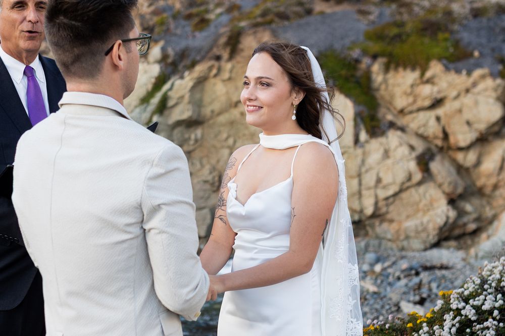 Bride in white gown and veil smiles at groom during outdoor coastal wedding ceremony with rocky cliffs backdrop.