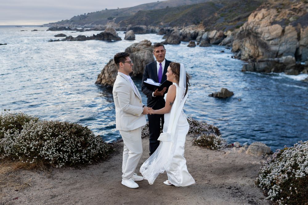 A romantic coastal wedding photoshoot with ocean waves crashing against rocky cliffs in the background.