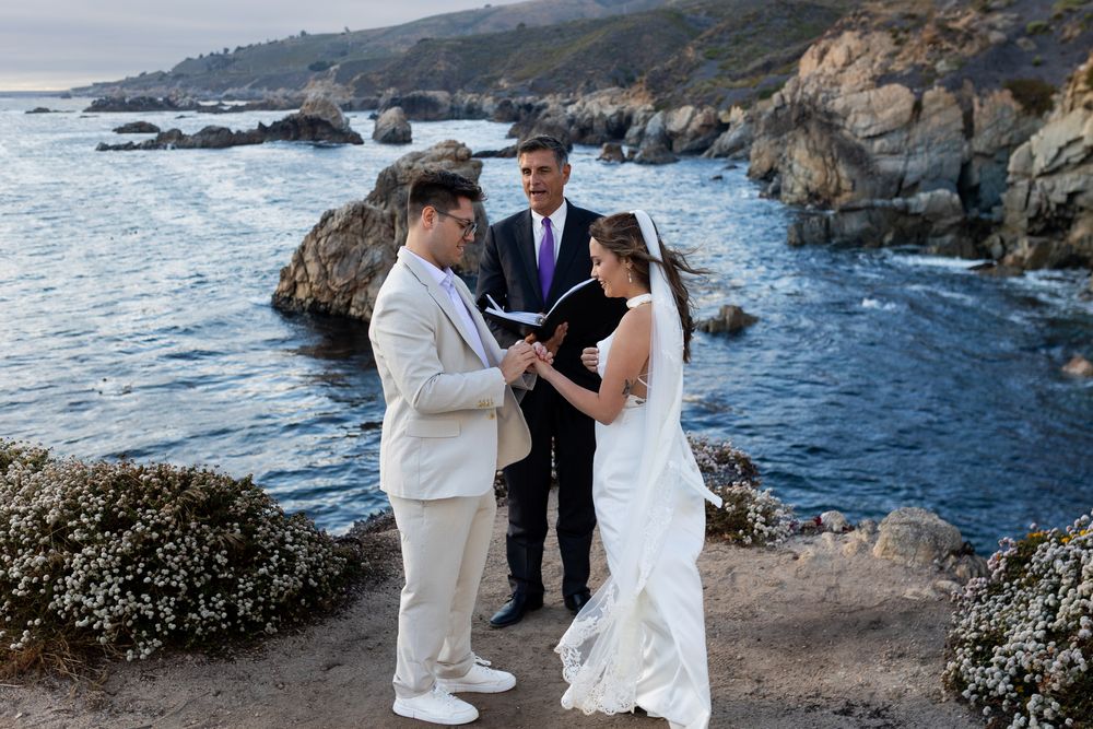 Couple exchanging vows at outdoor coastal wedding ceremony with rocky cliffs and ocean backdrop.