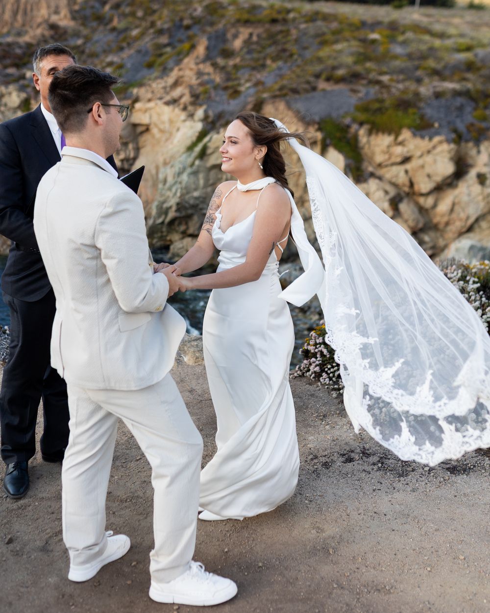 Bride and groom exchange vows outdoors near rocky cliffs, bride wearing white gown with lace veil, groom in cream suit.