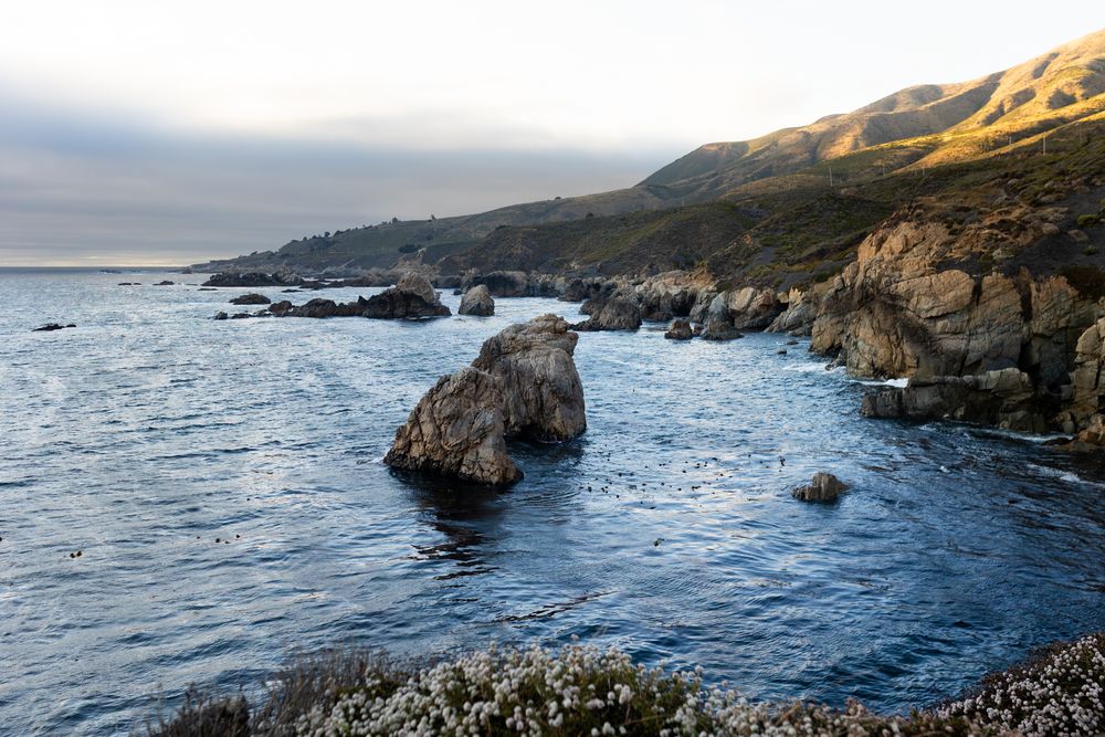 Scenic coastal landscape with rocky outcroppings emerging from blue ocean waters along a rugged hillside.