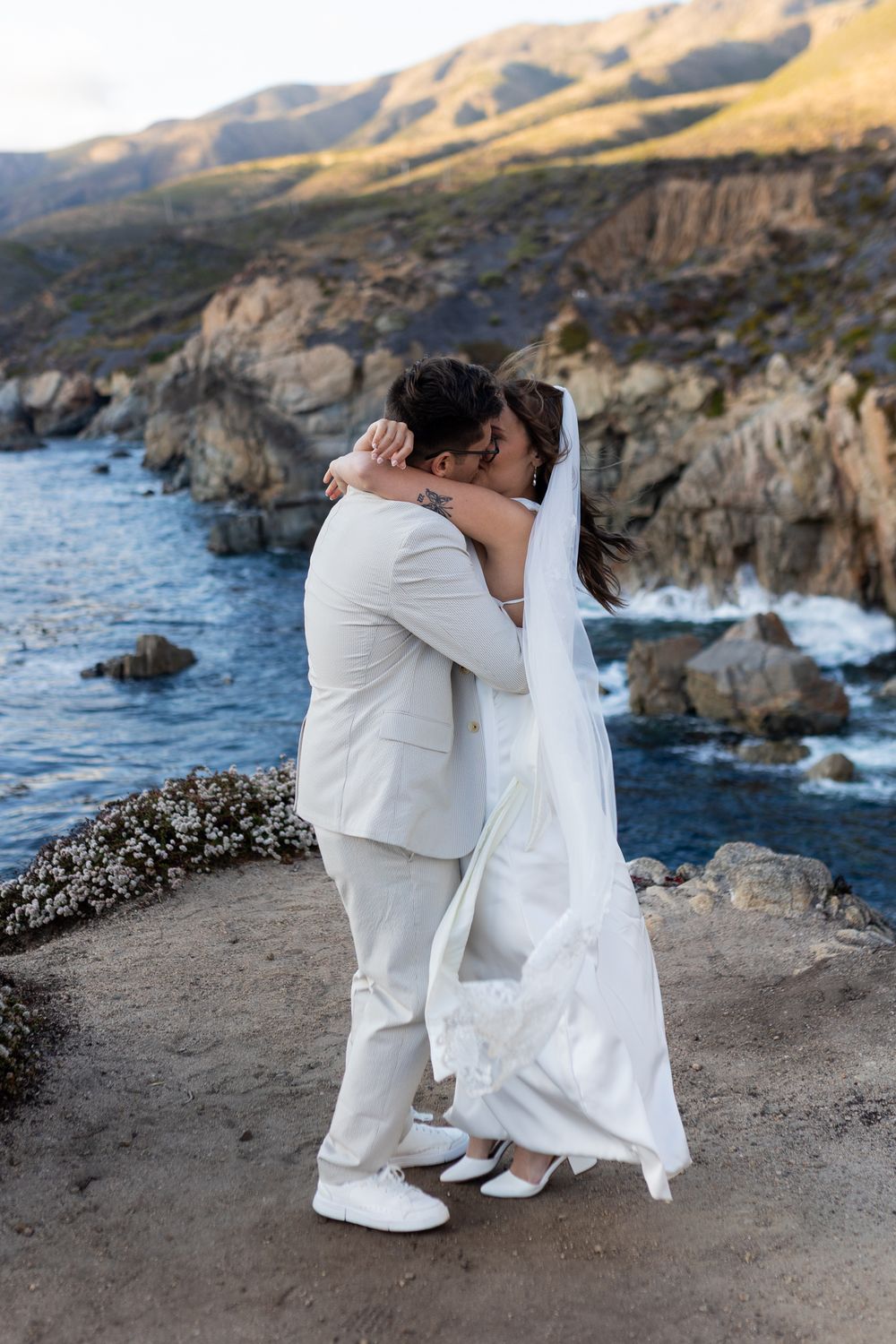 A couple in white attire shares an intimate moment on a scenic coastal cliff overlooking the ocean at sunset.